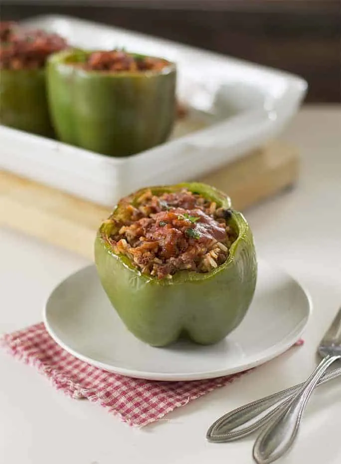 one green pepper stuffed with meat on a small white plate with a baking dish full of green stuffed peppers in the background on a wooden cutting board