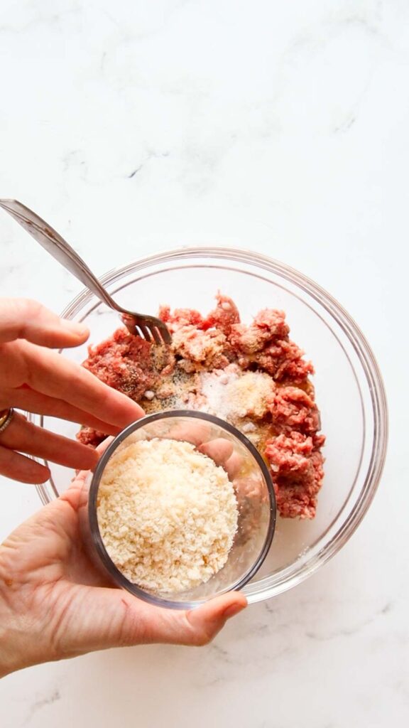 a hand showing bread crumbs in a bowl over ground beef 