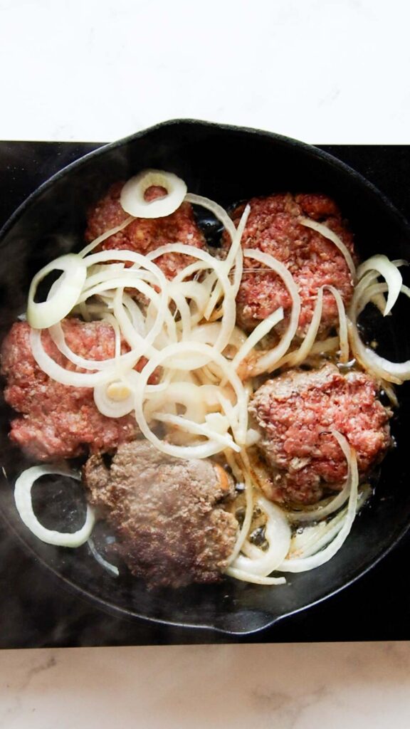 a skillet with cooking hamburger patties; onions on top 