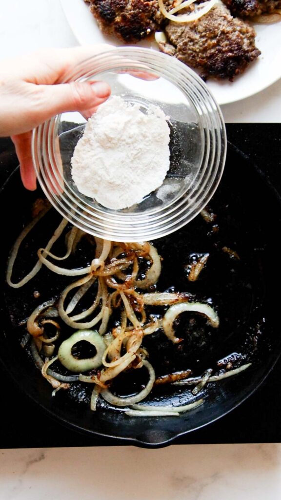 a hand adding flour in a bowl to a skillet with cooked onions 