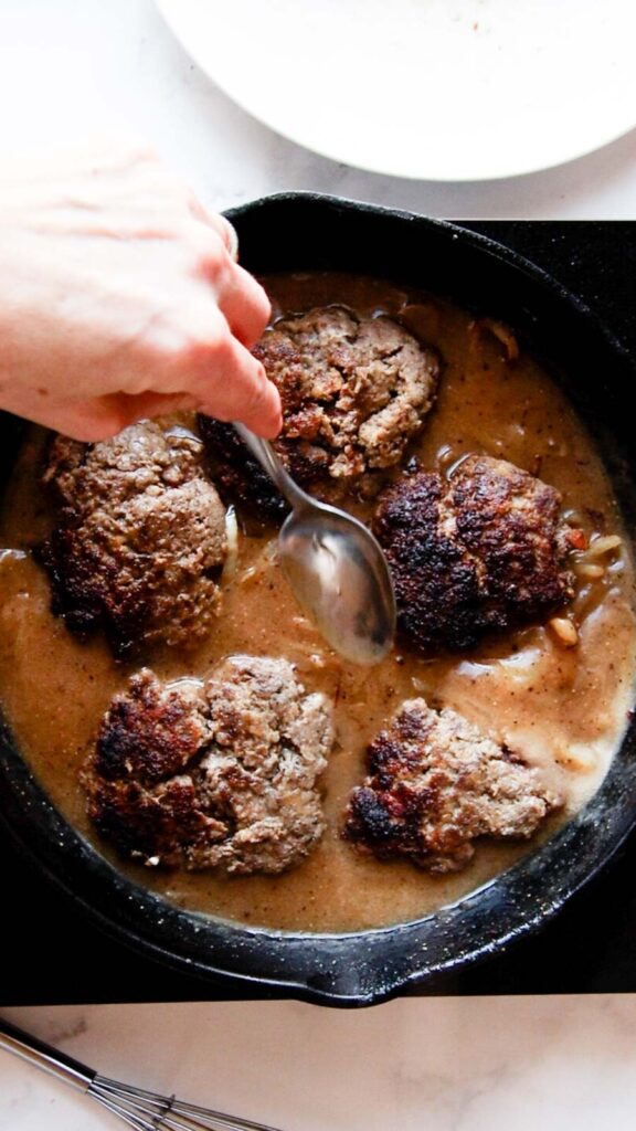 a skillet of gravy with hamburger steaks being added in to heat