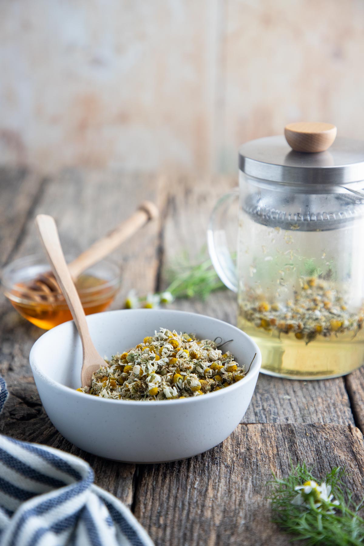 a bowl of dried chamomile flowers on a table with a spoon and teapot 