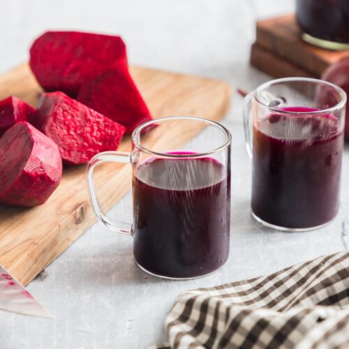 two glasses of beet kvass on a table with a cutting board with beets