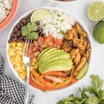 a chicken fajita bowl on a table and piled high with avocado, salsa, corn, beans, cilantro and rice
