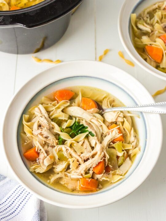 a bowl of chicken noodle soup with a spoon on a table and slow cooker in the background
