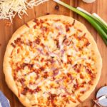 Overehead image of chicken bacon ranch pizza on a wooden work surface near cheese, scallions, and ranch dressing.