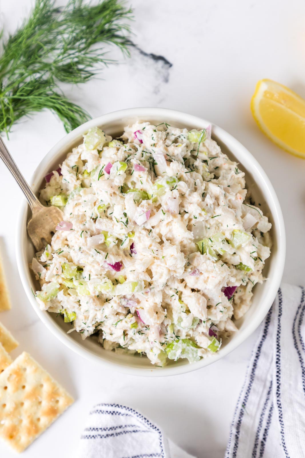 Overhead image of crab meat salad in a bowl near fresh dill, lemon, and crackers.