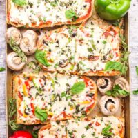 A pan of baked French bread pizzas with vegetables and fresh herbs surrounding them.