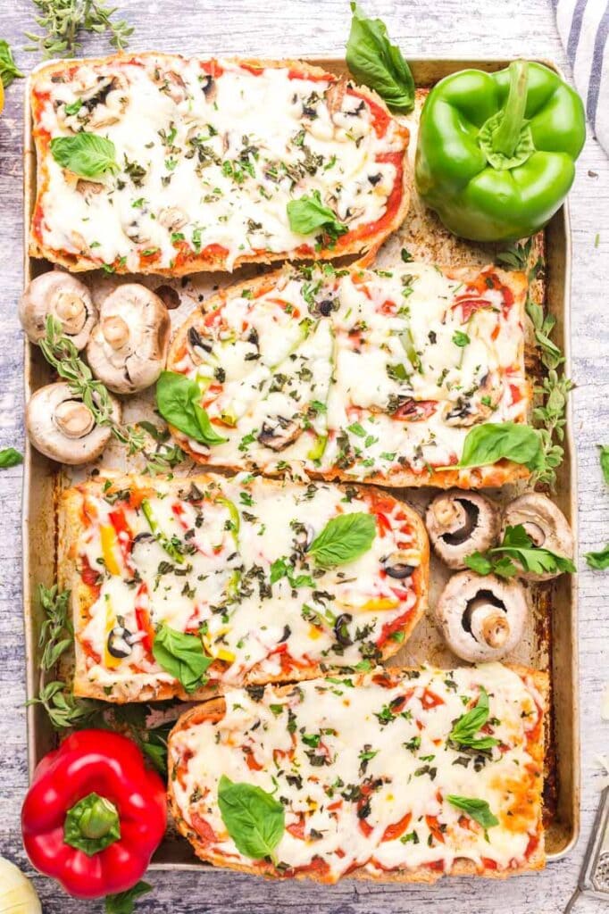 A pan of baked French bread pizzas with vegetables and fresh herbs surrounding them. 