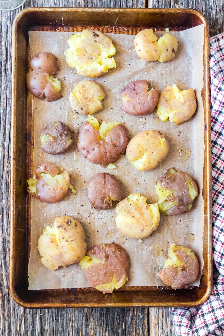 a baking tray with smashed potatoes that have avocado oil, salt and pepper