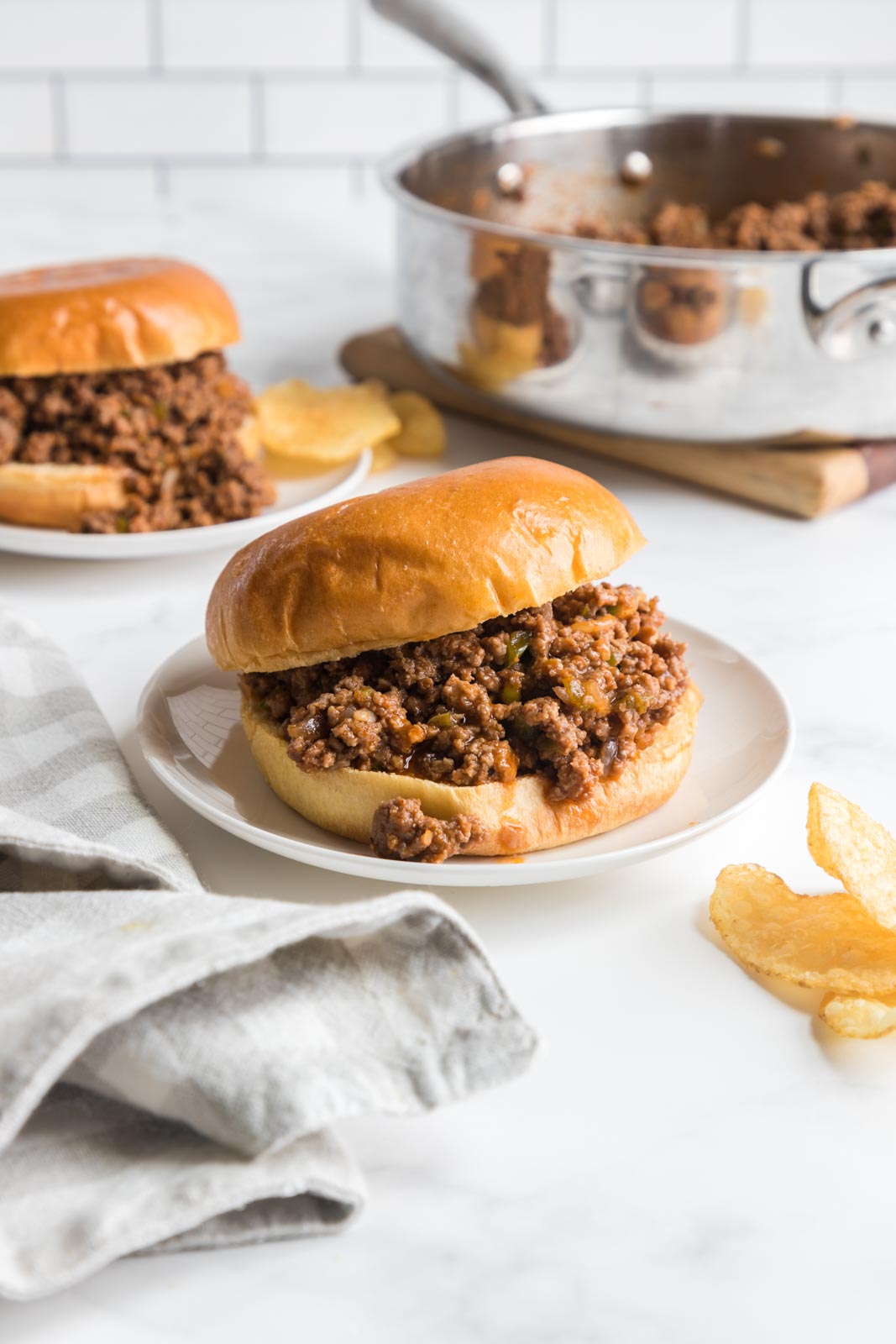 two sloppy joes on buns on plates on a table with a skillet in the background