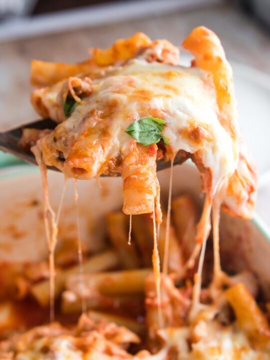 a spoonful of baked ziti being lifted from the pan on a spoon with strings of cheese