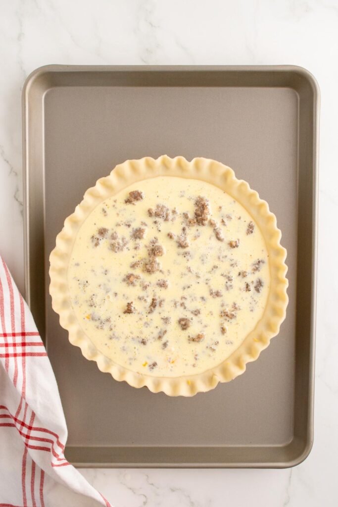 a pie crust with eggs poured over sausage and cheese ready to be baked on a cookie sheet