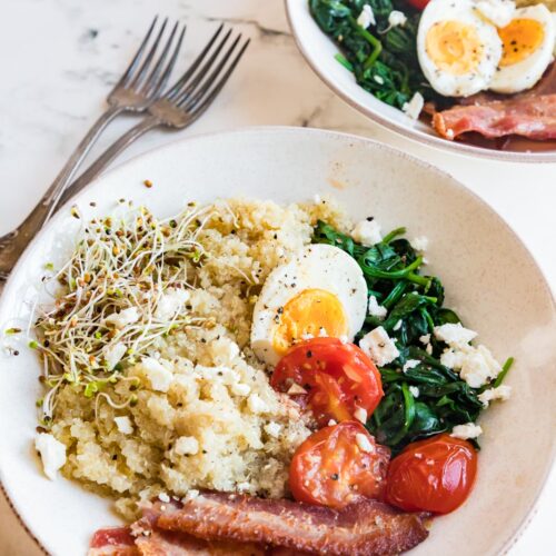 two breakfast bowls on a table with quinoa, eggs, bacon, sauteed spinach and tomatoes