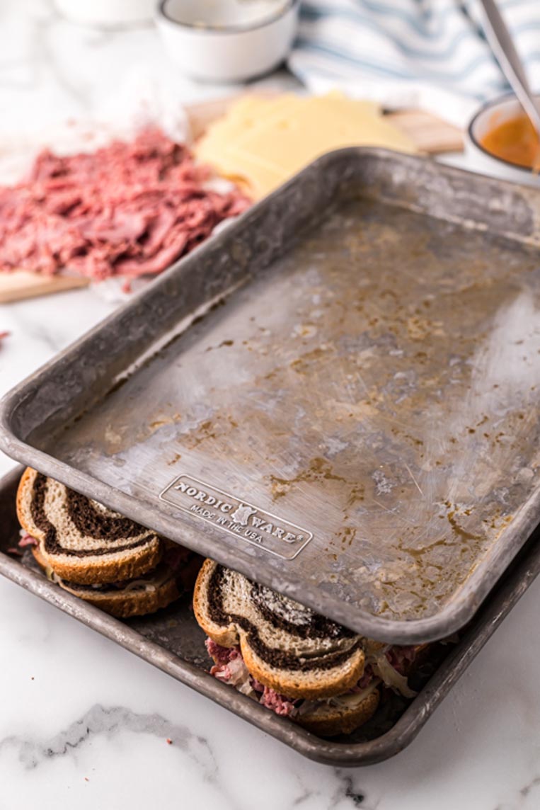 two cookie sheets on top of reuben sandwiches with them peeking out between