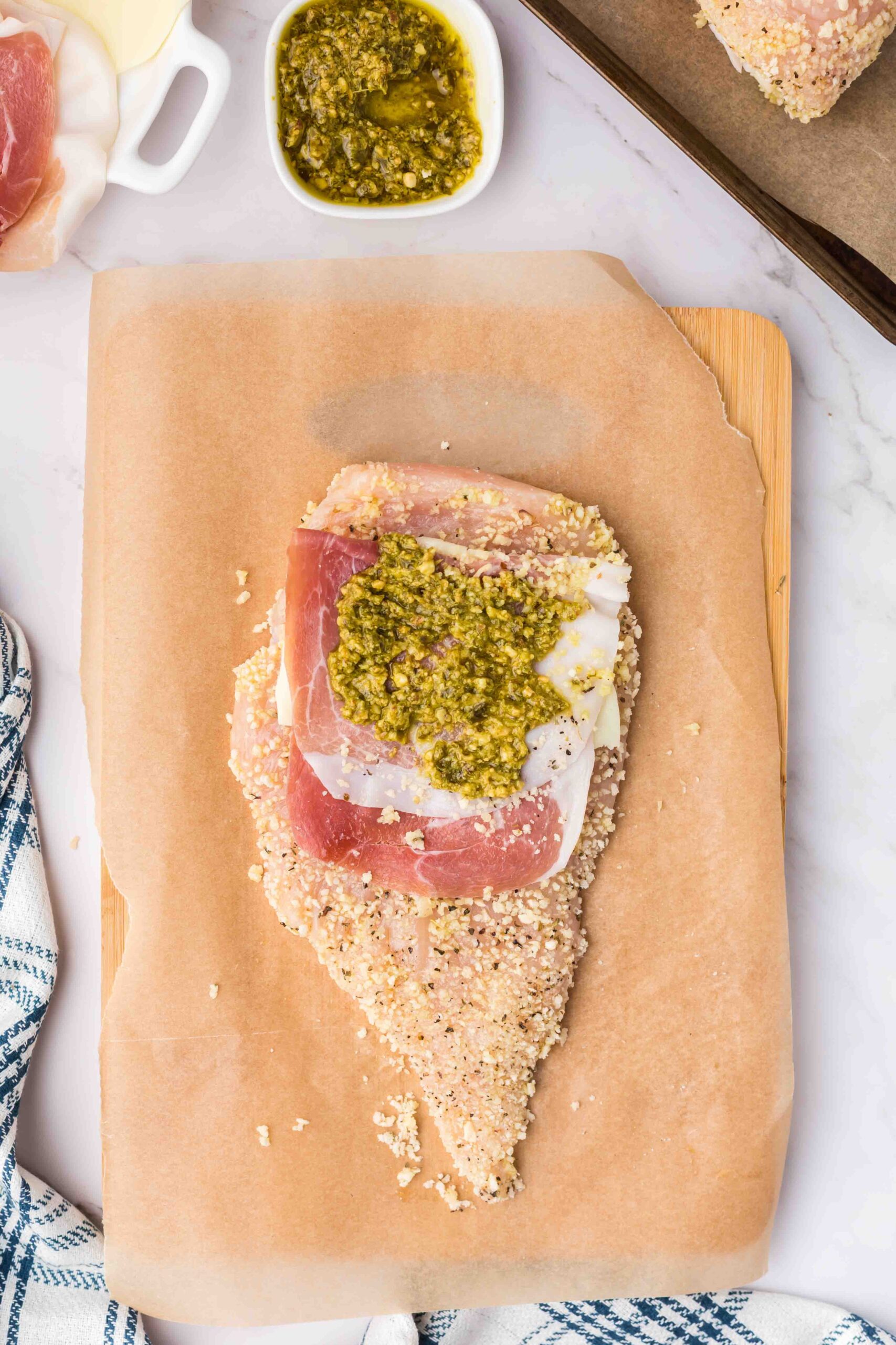 a chicken breast that's been breaded and a slice of cheese, prosciutto, and pesto on a table 