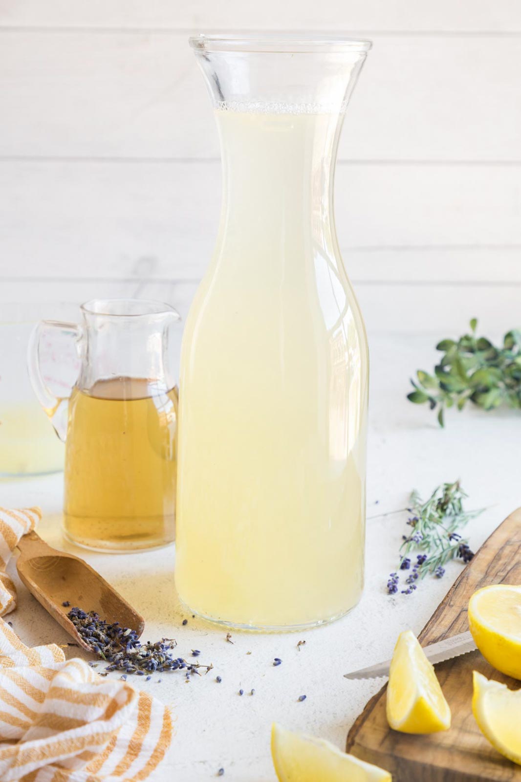 A pitcher of lavender lemonade near lemons, dried and fresh lavender, and lavender simple syrup.