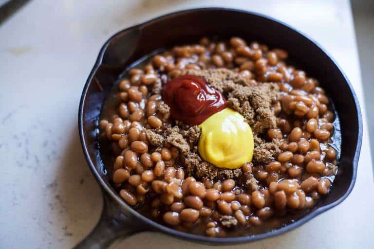 A skillet of baked beans with dollops of mustard, ketchup and brown sugar before being stirred together