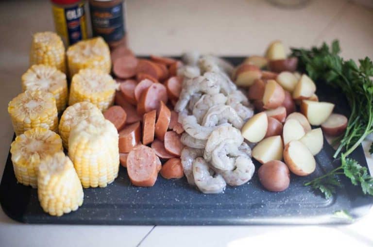 a cutting board with ingredients for shrimp boil packets; corn, sausage, shrimp, potatoes and herbs