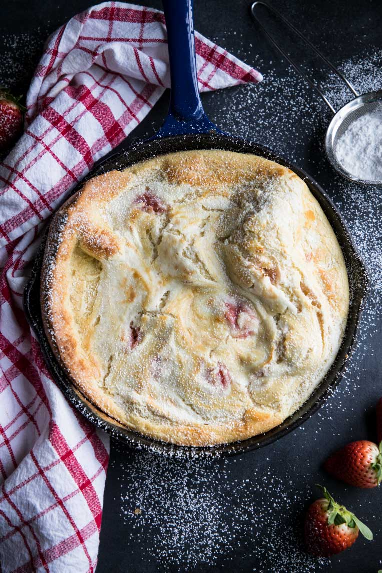 Dutch baby pancake in a skillet with powdered sugar and tomatoes. 