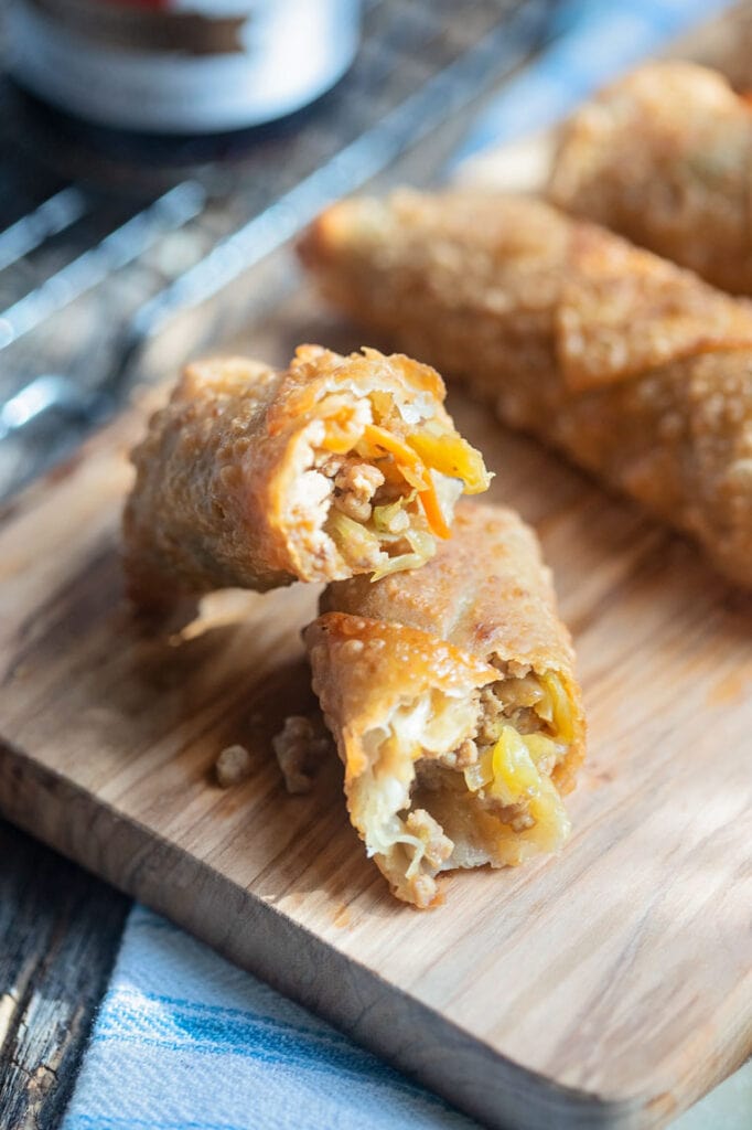 an egg roll broken in half  on a wooden cutting board with two egg rolls in the background on a blue napkin