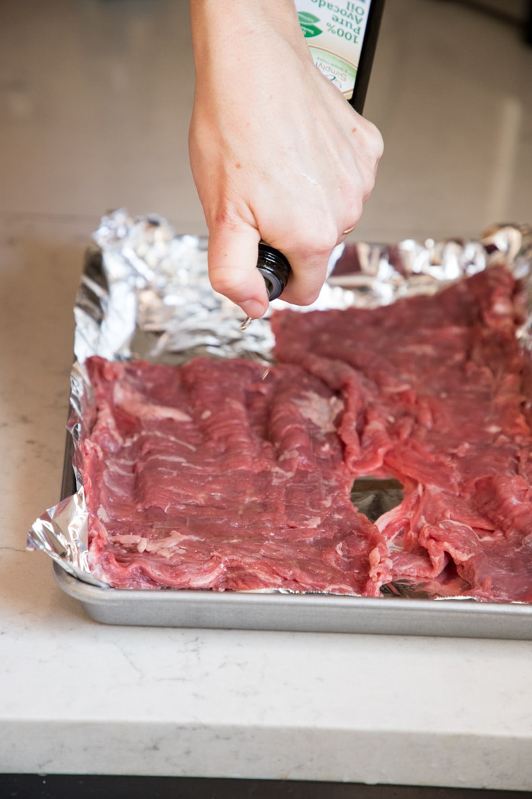 a person drizzling olive oil, vegetable oil or avocado oil on a flank or skirt steak on a tinfoil lined baking sheet