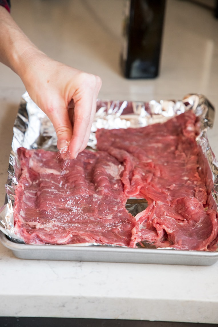 a person sprinkling salt and pepper on a flank or skirt steak on a foil lined baking sheet