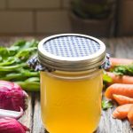 How to make homemade chicken stock. A jar of chicken stock on a table with vegetables.