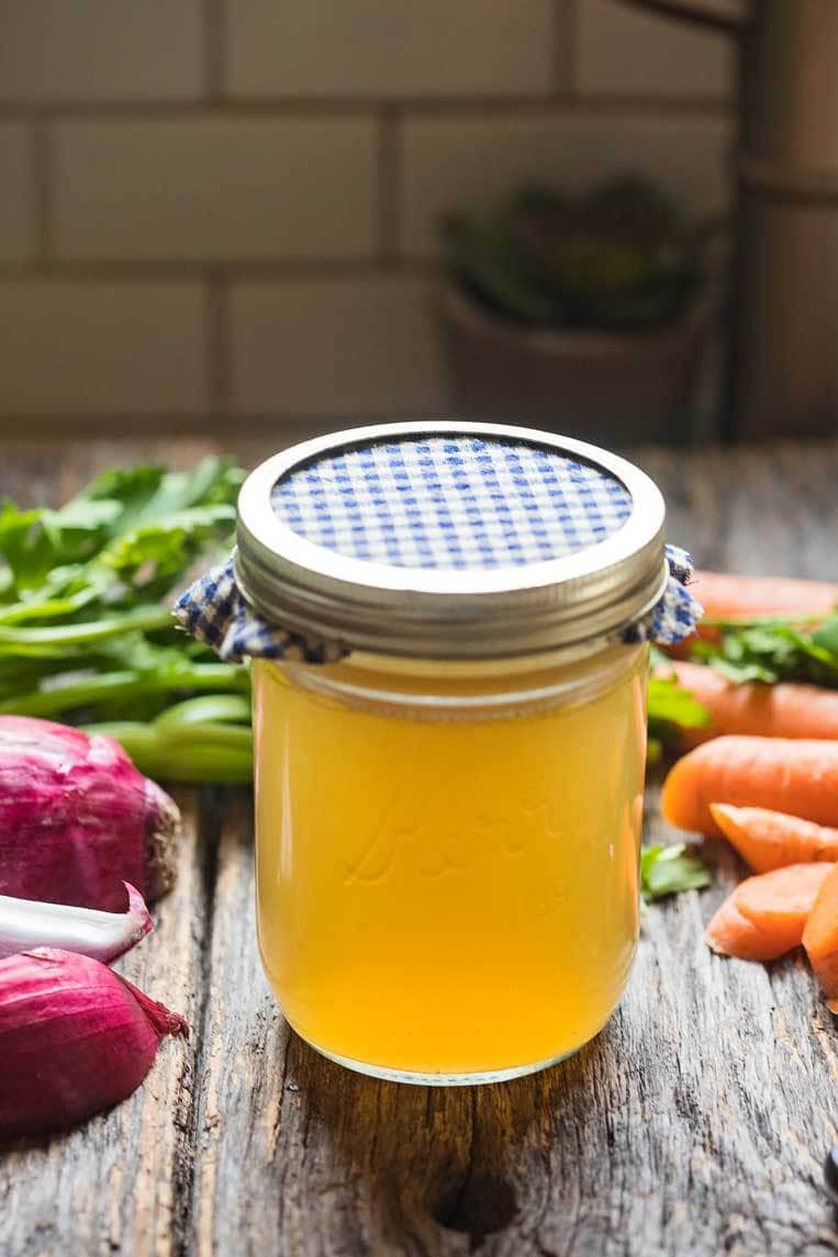 How to make homemade chicken stock. A jar of chicken stock on a table with vegetables.