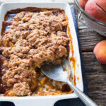 a big pan of fresh peach cobbler with a spoon in it by a window