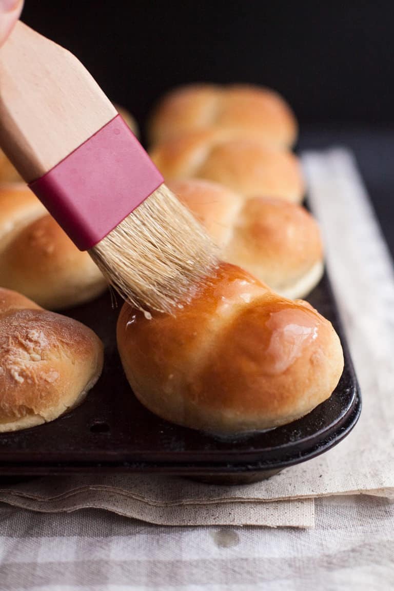 brush spreading warm butter over dinner rolls in a muffin tin